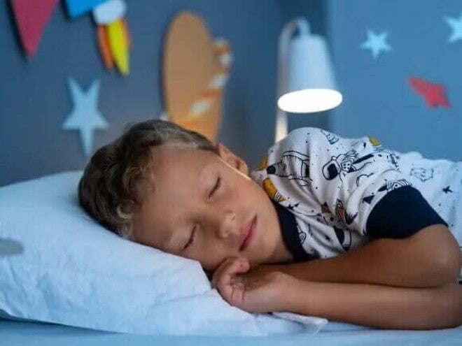 Criança dormindo Um menino, vestido de pijama estampado, está deitado na cama, de olhos fechados. O abajur atrás dele está aceso, e nas paredes do quarto há decorações de planetas.