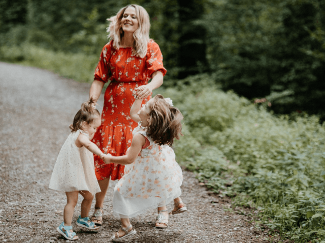 mulher, sorrindo e dançando com duas meninas pequenas em um campo, ambas vestindo vestidos brancos.