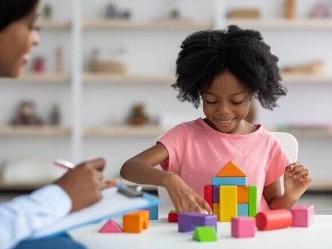 Uma menina sorrindo e organizando blocos coloridos, ao lado de uma mulher sentada com uma ficha na mão. Uma menina sorrindo e organizando blocos coloridos, ao lado de uma mulher sentada com uma ficha na mão.