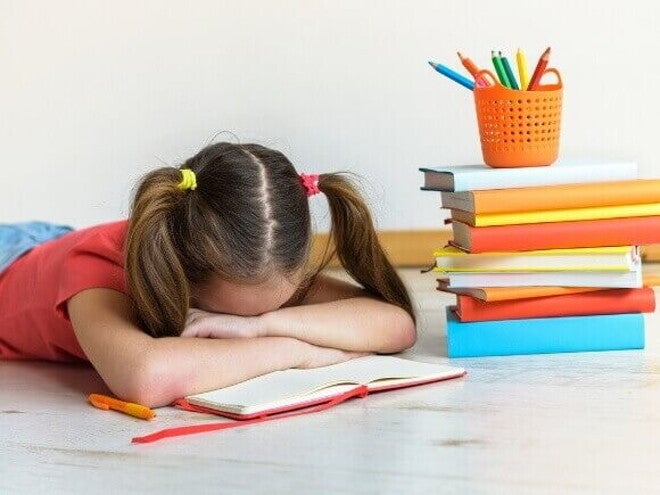 Menina deitada sobre um caderno aberto com a cabeça entre os braços, demonstrando exaustão e dificuldade de aprendizagem diante de uma pilha de livros e materiais escolares.