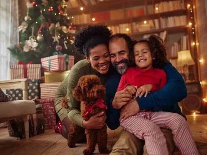 Homem e mulher abraçados com a filha e um cachorrinho no chão, na sala de casa em frente a uma árvore de natal Homem e mulher abraçados com a filha e um cachorrinho no chão, na sala de casa em frente a uma árvore de natal