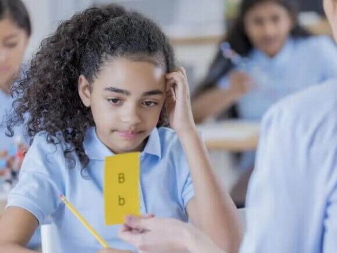 Dislexia infantil Imagem de uma criança com dislexia olhando para uma ficha amarela com letras B maiúscula e minúscula. Em uma mão ela segura um lápis e a outra está sobre a cabeça em uma postura de dúvida.