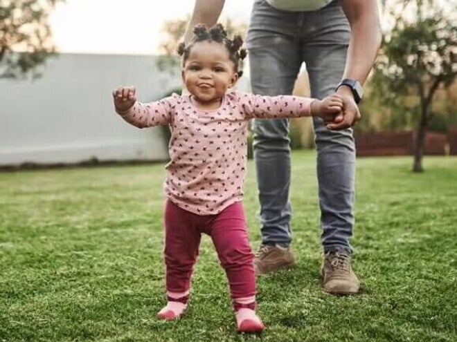 Imagem de um bebê cuja criação segue a abordagem de Emmi Pikler e que está brincando em um quintal supervisionado pela mãe que está lhe dando a mão. Imagem de um bebê cuja criação segue a abordagem de Emmi Pikler e que está brincando em um quintal supervisionado pela mãe que está lhe dando a mão.