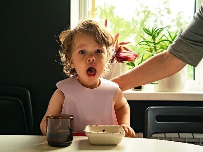 Um bebê com babador sentado à mesa, com a boca aberta, olhando diretamente para a câmera, com a mão de um adulto ao fundo. Um bebê com babador sentado à mesa, com a boca aberta, olhando diretamente para a câmera, com a mão de um adulto ao fundo.
