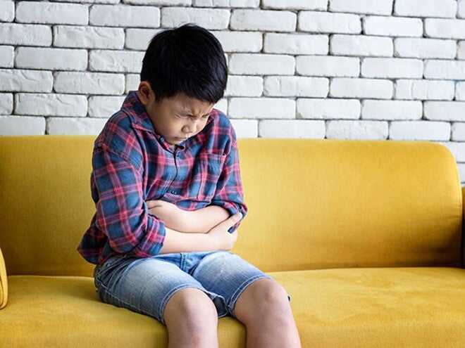 Estomatite infantil: o que é? Causas, sintomas e tratamento menino sentado no sofá com feição de dor abraçando a barriga