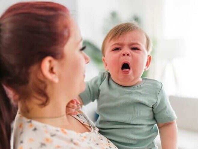 Mãe segurando um bebê que chora, com a boca aberta, indicando dor ou desconforto, possivelmente devido à faringite em bebês ou outra inflamação na garganta.
