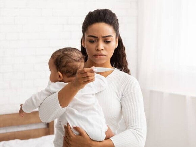 Uma mulher segura um bebê nos braços, com feição preocupada, enquanto verifica a temperatura do bebê com um termômetro.