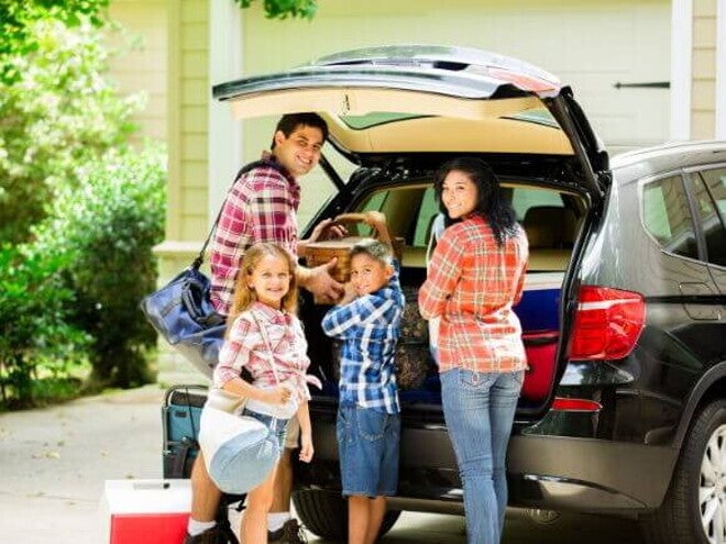 Família posando e sorrindo para a câmera, enquanto organiza malas dentro de um carro preto para sair de viagem Família posando e sorrindo para a câmera, enquanto organiza malas dentro de um carro preto para sair de viagem