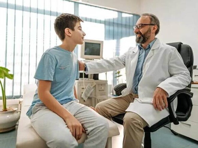 Imagem de um ambiente hospitalar em que um médico tranquiliza um paciente menino, ao colocar a mão no ombro dele. O médico está de jaleco branco, óculos e tem barba e o menino está vestindo uma camiseta azul Imagem de um ambiente hospitalar em que um médico tranquiliza um paciente menino, ao colocar a mão no ombro dele. O médico está de jaleco branco, óculos e tem barba e o menino está vestindo uma camiseta azul