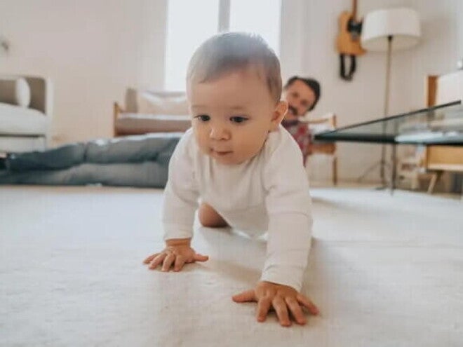 Bebê engatinhando no chão da sala de casa, com a supervisão do pai olhando do canto da sala