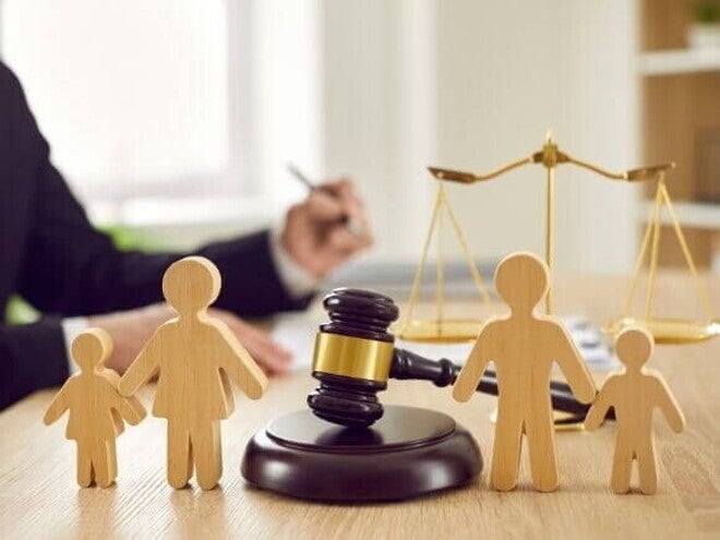 Objetos de direito sobre uma mesa de madeira, incluindo um martelo de juiz e bonecos de madeira representando uma família. A balança da justiça aparece ao fundo, e a imagem, com um juiz escrevendo ao fundo, reflete um processo judicial para a decisão de como funciona a guarda unilateral. Objetos de direito sobre uma mesa de madeira, incluindo um martelo de juiz e bonecos de madeira representando uma família. A balança da justiça aparece ao fundo, e a imagem, com um juiz escrevendo ao fundo, reflete um processo judicial para a decisão de como funciona a guarda unilateral.