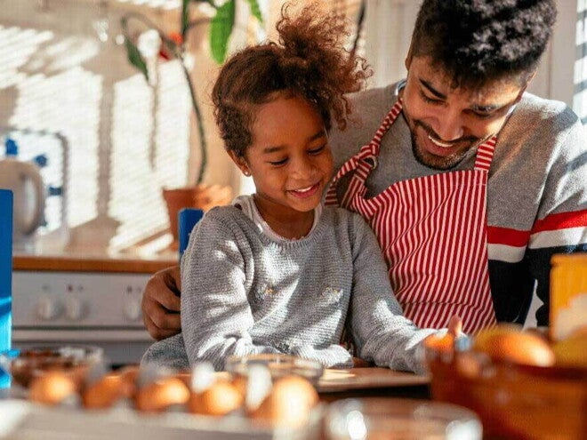 A relação de pai e filho é ilustrada por uma imagem onde a menina é abraçada pelo pai, que veste avental, enquanto eles olham para uma mesa de café da manhã com alguns alimentos.