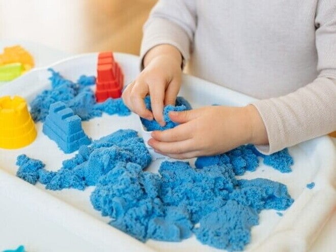Close das mãos de uma criança interagindo com areia cinética azul em uma bandeja branca.