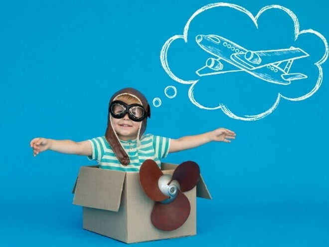 Um menino usando óculos de aviador e um capacete de couro decola de dentro de uma caixa de papelão, sorrindo. Acima de sua cabeça, uma bolha de pensamento branca sobre um fundo azul vibrante mostra um desenho de um avião.