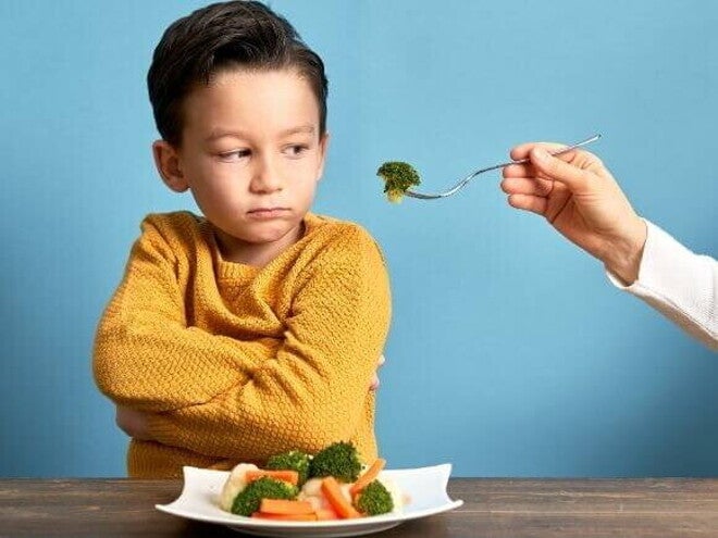 Uma mão estendendo um garfo com brócolis para um menino, que olha de forma irritada para o alimento. Uma mão estendendo um garfo com brócolis para um menino, que olha de forma irritada para o alimento.