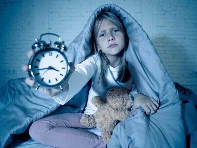 Uma menina loira, com uma expressão de cansaço ou preocupação, sentada na cama coberta por um edredom, segurando um despertador à sua frente, com um ursinho de pelúcia ao lado.