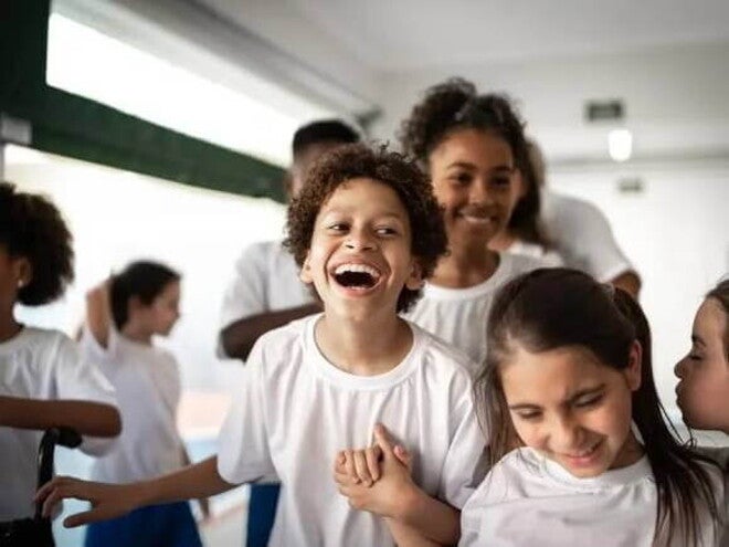 Imagem de um ambiente escolar com várias crianças vestidas de uniforme. O destaque está para um menino gargalhando, bem no centro da foto, de mãos dadas com uma menina Imagem de um ambiente escolar com várias crianças vestidas de uniforme. O destaque está para um menino gargalhando, bem no centro da foto, de mãos dadas com uma menina
