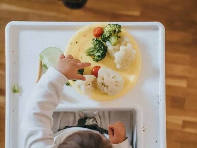 Introdução alimentar Uma fotografia tirada de cima mostra um bebê em fase de introdução alimentar e seu prato amarelo com vegetais coloridos a sua frente