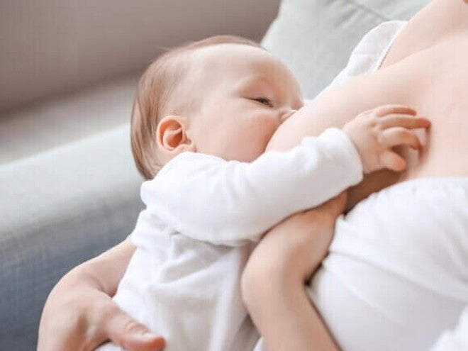 Bebê de roupa branca sendo amamentado pela mãe sentada em um sofá Bebê de roupa branca sendo amamentado pela mãe sentada em um sofá
