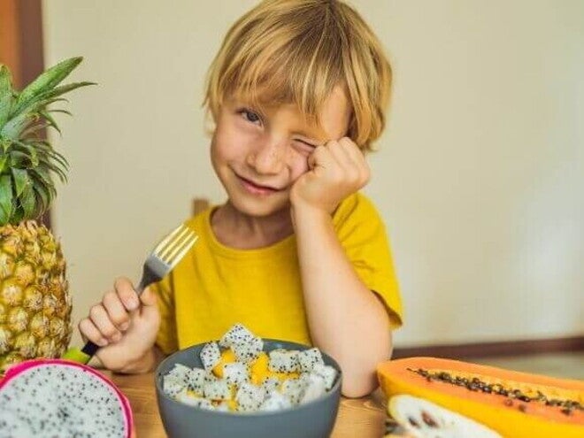 Um menino sentado à mesa com um prato de frutas variadas segurando um garfo e piscando um olho. A imagem representa um lanche da tarde para crianças com frutas saudáveis. Um menino sentado à mesa com um prato de frutas variadas segurando um garfo e piscando um olho. A imagem representa um lanche da tarde para crianças com frutas saudáveis.