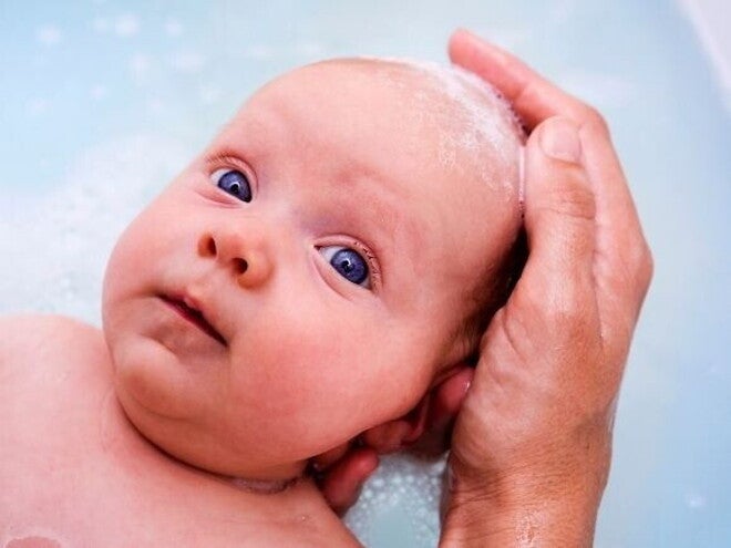 Um close-up de um bebê recém-nascido, deitado na água do banho com espuma. Uma mão adulta com sabão na ponta dos dedos está suavemente lavando a cabeça do bebê.