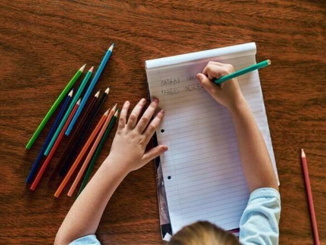 Um menino está escrevendo em um bloco de folhas alguns nomes, como “Fabio”. Na mesa estão espalhados lápis de cor de diversas cores. A foto está ligada ao tema Troca de letras na infância. Um menino está escrevendo em um bloco de folhas alguns nomes, como “Fabio”. Na mesa estão espalhados lápis de cor de diversas cores. A foto está ligada ao tema Troca de letras na infância.