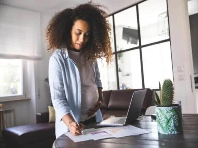 Uma mulher grávida escreve em alguns papéis na mesa da sala de casa, enquanto está de licença-maternidade. Uma mulher grávida escreve em alguns papéis na mesa da sala de casa, enquanto está de licença-maternidade.