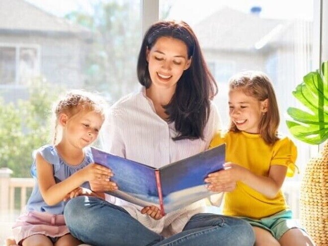 Uma mulher sorridente está sentada lendo um livro para duas meninas pequenas sentadas ao seu lado. Uma mulher sorridente está sentada lendo um livro para duas meninas pequenas sentadas ao seu lado.