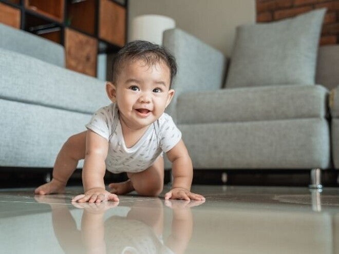 Um bebê sorridente engatinhando em um piso brilhante, com um sofá cinza e uma parede de tijolos ao fundo, ilustrando um marco de desenvolvimento motor.