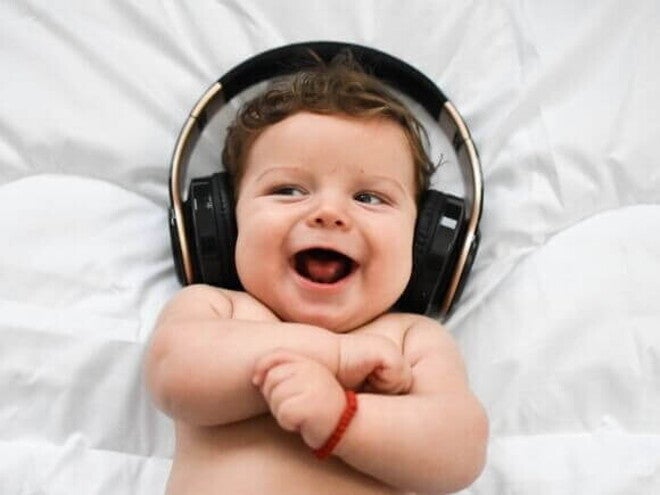 Música para bebês Um bebê sorridente, deitado em uma superfície branca, usa fones de ouvido grandes e pretos sobre a cabeça.