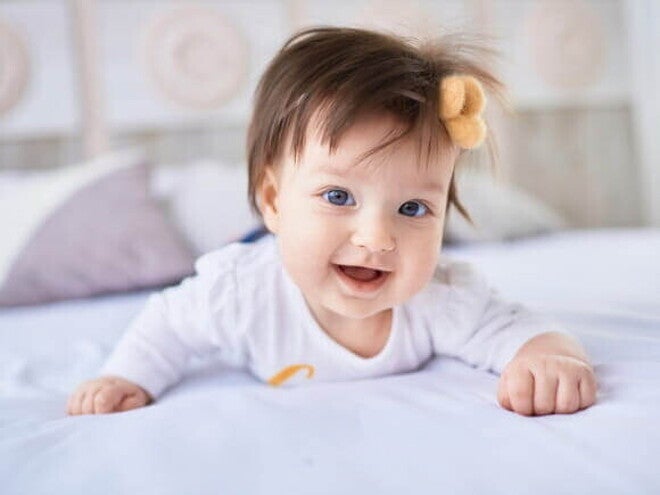 Bebê sorridente deitada de bruços na cama, usando roupa branca e um laço no cabelo Bebê sorridente deitada de bruços na cama, usando roupa branca e um laço no cabelo