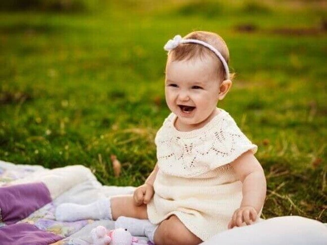 Bebê sorridente usando um vestido claro e uma tiara com flores, sentada em um cobertor sobre a grama. Bebê sorridente usando um vestido claro e uma tiara com flores, sentada em um cobertor sobre a grama.