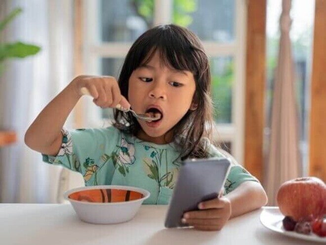 Uma menina come enquanto olha para um celular na mesa. Uma menina come enquanto olha para um celular na mesa.