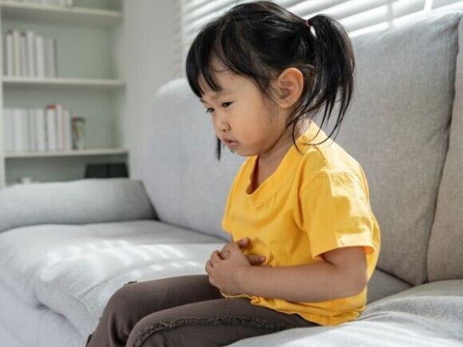 Uma menina está sentada em um sofá, segurando a barriga com as duas mãos, com uma expressão de desconforto ou dor. Uma menina está sentada em um sofá, segurando a barriga com as duas mãos, com uma expressão de desconforto ou dor.