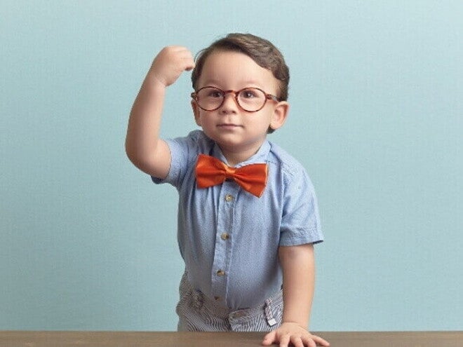 Menino com óculos, gravata vermelha e camisa azul, está apoiado sobre uma mesa, com a mão direita levantada em punho, sob um fundo azul. Menino com óculos, gravata vermelha e camisa azul, está apoiado sobre uma mesa, com a mão direita levantada em punho, sob um fundo azul.