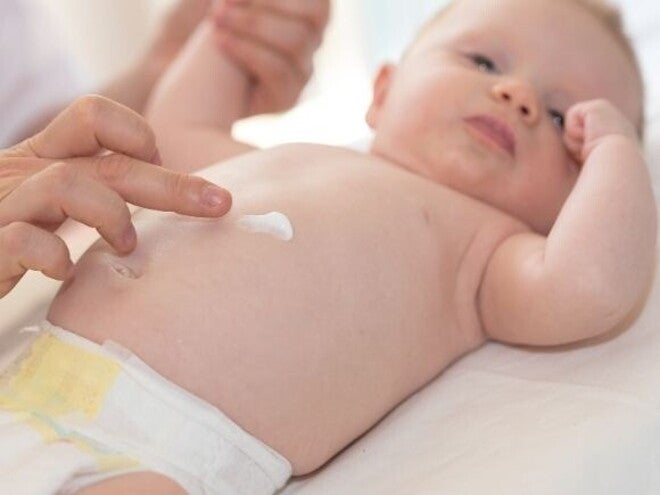 Uma mão de adulto gentilmente aplica creme na barriga de um bebê deitado, destacando o cuidado com a pele delicada do bebê.