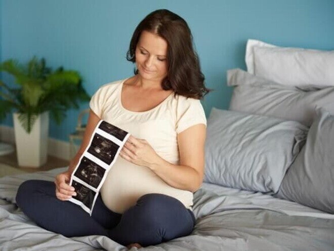 Mulher grávida sentada em uma cama, observando o resultado de um exame de ultrassom Mulher grávida sentada em uma cama, observando o resultado de um exame de ultrassom