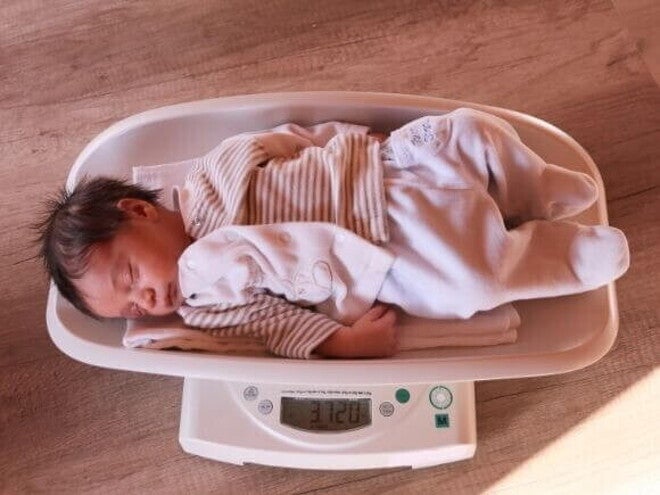 Um bebê recém-nascido dormindo sobre uma balança digital que mostra o peso. A imagem ilustra o monitoramento do peso do bebê, um tema central para a perda de peso neonatal.