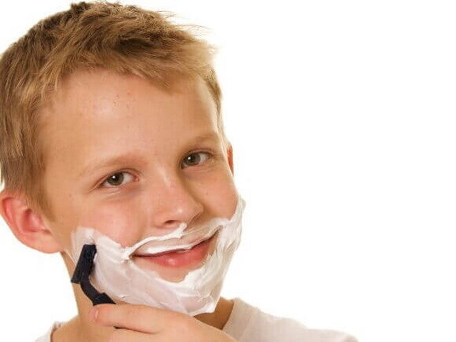 Um menino passando com creme de barbear no rosto, passando uma lâmina de barbear na bochecha e sorrindo Um menino passando com creme de barbear no rosto, passando uma lâmina de barbear na bochecha e sorrindo