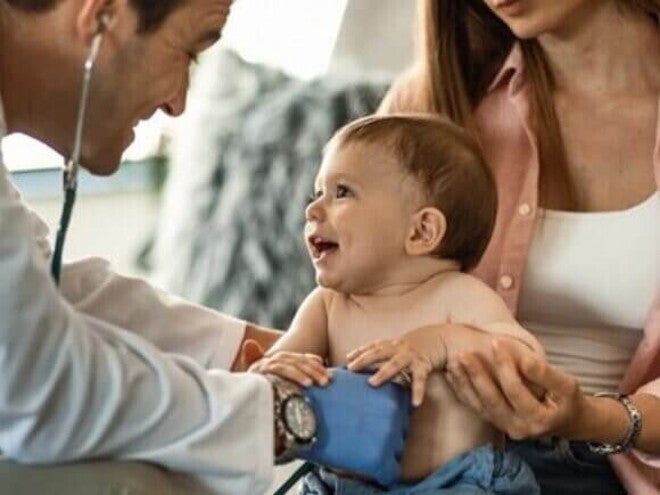 Menino sorridente de aproximadamente um ano sentado no colo da mãe recebendo atendimento médico Menino sorridente de aproximadamente um ano sentado no colo da mãe recebendo atendimento médico