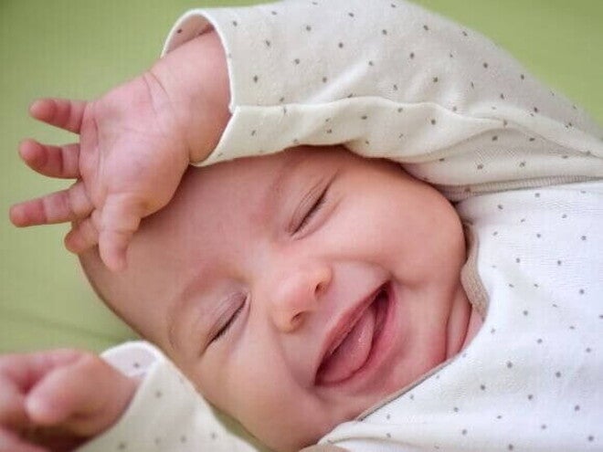 Close de um bebê recém-nascido deitado, com os olhos fechados e uma expressão de felicidade. Ele usa um macacão branco com bolinhas pretas e está com a mãozinha sobre a testa.