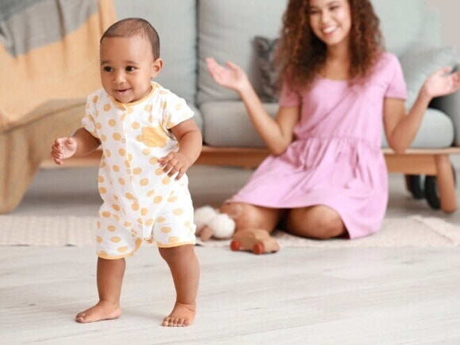 Um bebê sorrindo e em pé no chão, olhando para a frente, com uma mulher sentada no chão ao fundo, sorrindo. Um bebê sorrindo e em pé no chão, olhando para a frente, com uma mulher sentada no chão ao fundo, sorrindo.