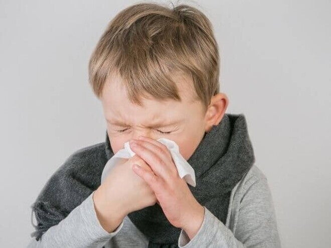 Um menino segurando um lenço de papel para assoar o nariz. Ele está com os olhos fechados e o rosto contraído, demonstrando sintomas de resfriado em crianças, como congestão nasal e coriza. Um menino segurando um lenço de papel para assoar o nariz. Ele está com os olhos fechados e o rosto contraído, demonstrando sintomas de resfriado em crianças, como congestão nasal e coriza.