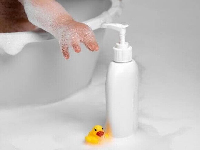 Mão de um bebê se esticando para alcançar um frasco de shampoo ao lado de um pato de borracha. Mão de um bebê se esticando para alcançar um frasco de shampoo ao lado de um pato de borracha.