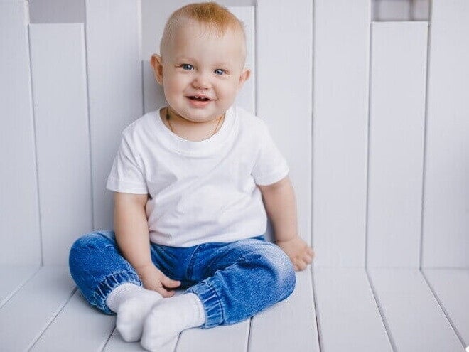 Um bebê sentado, sorrindo, veste uma camisa branca e uma calça jeans, sob um fundo branco. Um bebê sentado, sorrindo, veste uma camisa branca e uma calça jeans, sob um fundo branco.