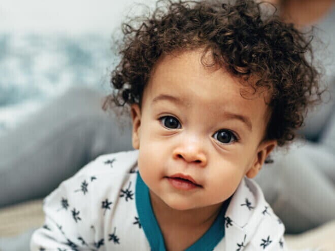 Bebê com cabelo cacheado vestindo um pijama branco com detalhes azuis, olhando diretamente para a câmera. Bebê com cabelo cacheado vestindo um pijama branco com detalhes azuis, olhando diretamente para a câmera.