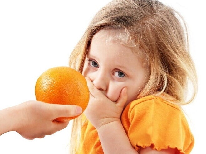 Uma mão oferece uma laranja a uma menina, que cobre a boca com a mão e olha para a câmera com uma expressão de repulsa ou aversão.