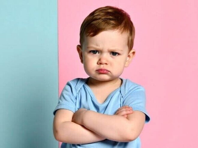 Fotografia de um menino pequeno expressando teimosia infantil. Ele está de braços cruzados, com uma expressão facial fechada e lábios comprimidos, vestindo uma camiseta azul contra um fundo dividido entre azul claro e rosa.