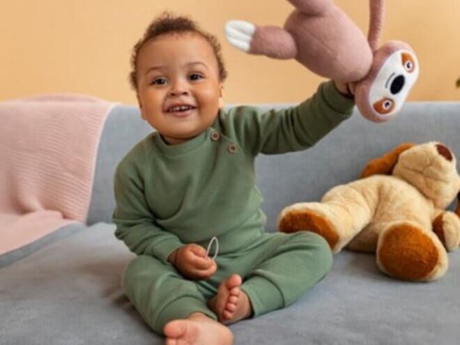 Menino usando macacão verde sentado no sofá segurando ursinho de pelúcia e sorrindo Menino usando macacão verde sentado no sofá segurando ursinho de pelúcia e sorrindo