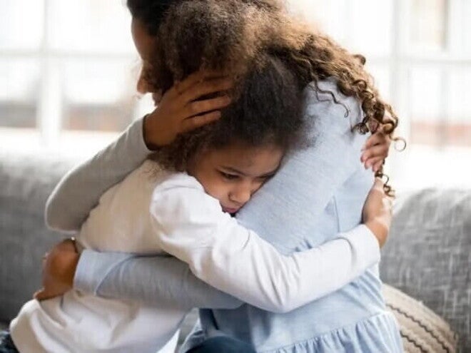 TOD Sentadas em um sofá cinza, uma mulher, usando uma blusa azul clara, abraça uma menina, que usa uma blusa branca. A menina está triste.